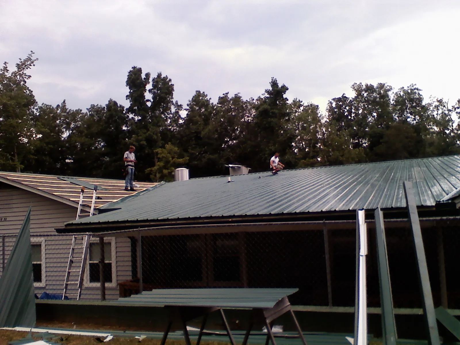 Two roofers installing green metal panels above a covered porch in Clearfield